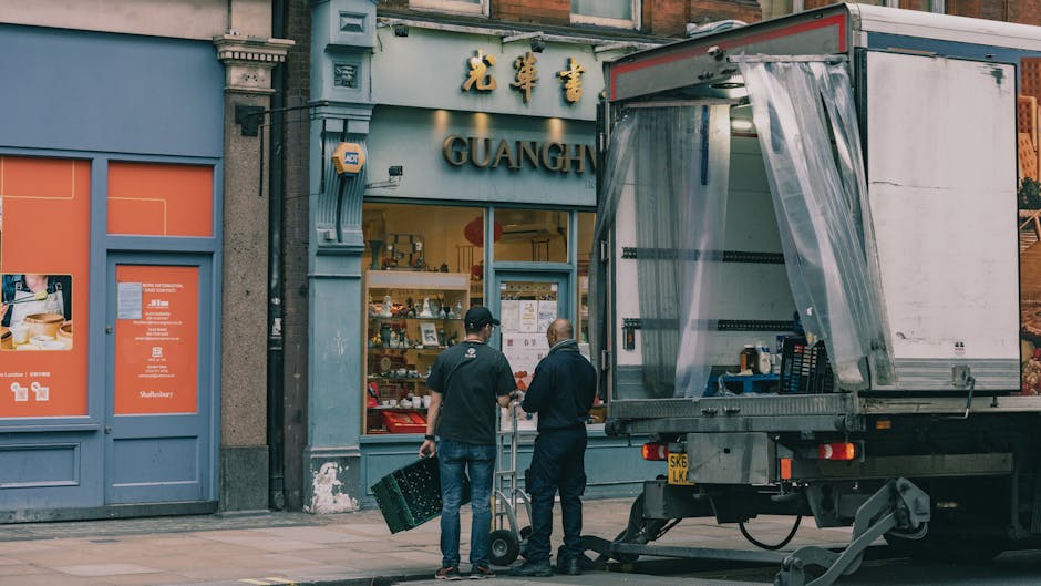 A wooden and metal moving van with an open side door parked on a city street in Hampstead, adjacent to a red British telephone booth. The van's interior contains a blue hand truck, yellow straps, and 