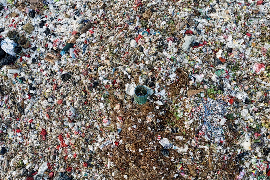 An aerial view of a large rubbish dump containing a mix of waste materials including plastic bottles, cardboard boxes, crumpled paper, fabric scraps, and discarded food packaging, covering the ground