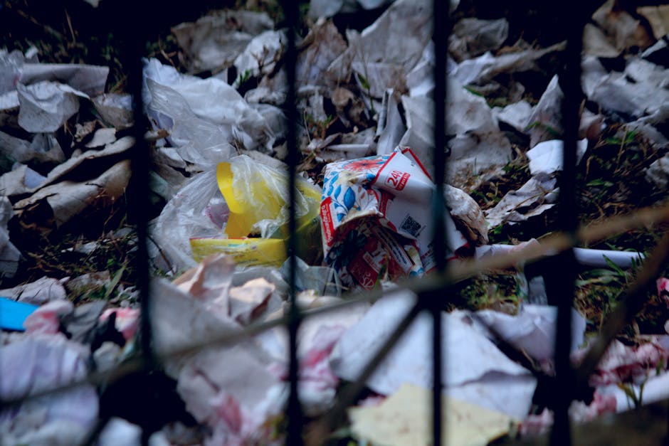 An aerial view of a large rubbish dump containing a mix of waste materials including plastic bottles, cardboard boxes, crumpled paper, fabric scraps, and discarded food packaging, covering the ground