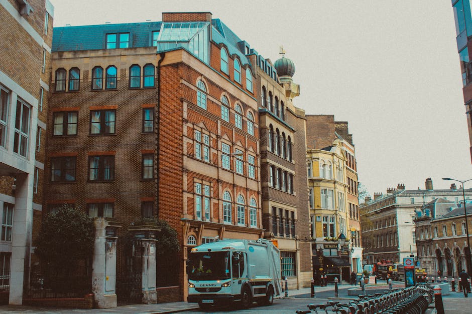 A small maroon flatbed truck parked on a city street in Hampstead, loaded with various packed items including black and white plastic bags, wrapped furniture, and cardboard boxes, all secured with str
