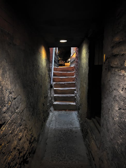 A three-flight metal fire escape staircase with grated steps and railings, attached to the exterior of a multi-storey brick building in Hampstead. The staircase is positioned inside a narrow alleyway 