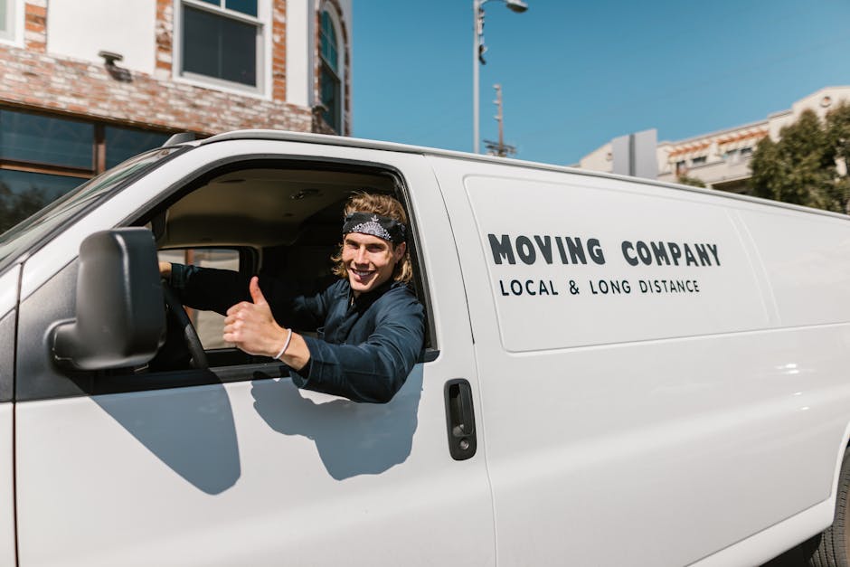 A young male mover with long hair, wearing a black bandana and navy blue shirt, sitting in the driver's seat of a white moving van. The side of the van displays the text 'MOVING COMPANY' and 'LOCAL &